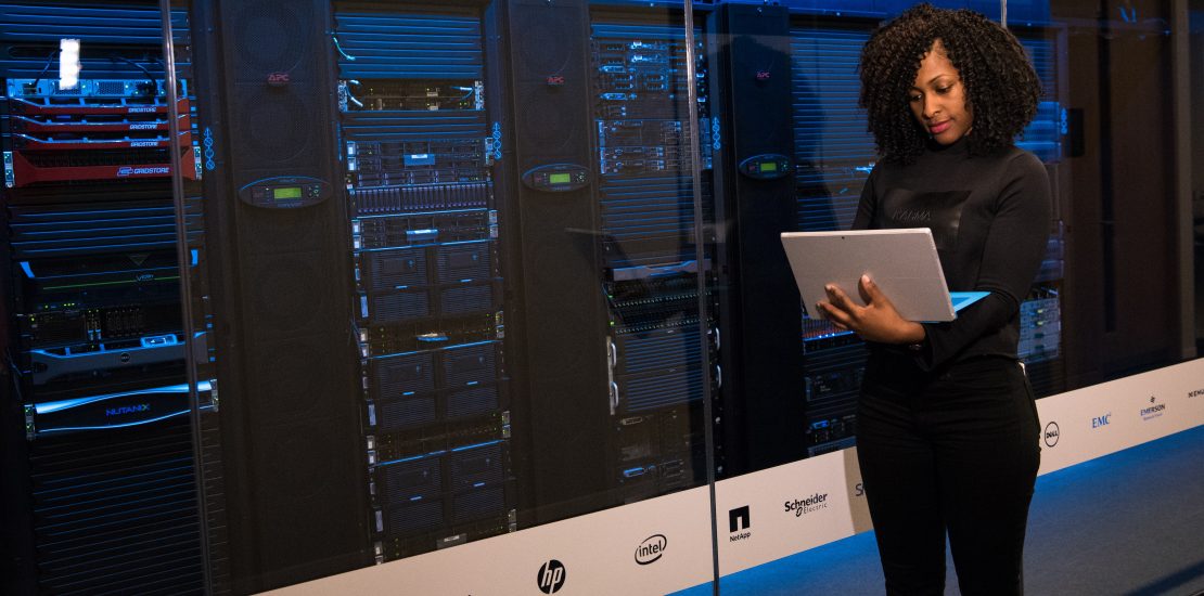 Woman using a laptop while standing in front of multiple mainframe racks